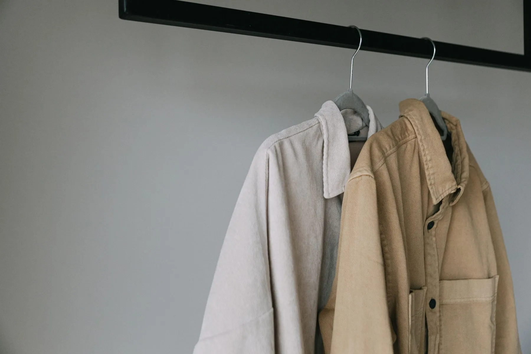 Dos camisas gruesas colgadas, una en crudo de pana y otra en camel, sobre fondo gris