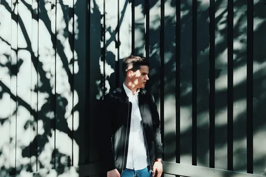 Hombre con chaqueta bomber negra, camisa blanca y vaqueros azules, apoyado en una valla con sombras de ramas