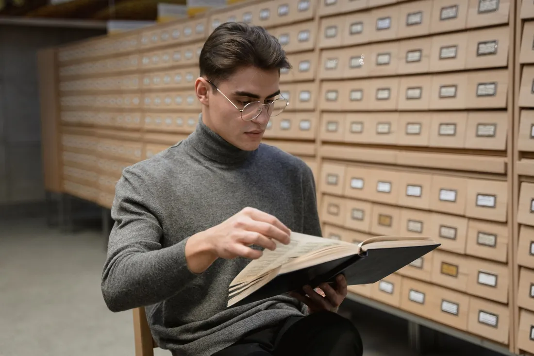 Hombre con gafas redondas y jersey de cuello vuelto gris leyendo un libro sentado junto a un mueble de archivo de madera