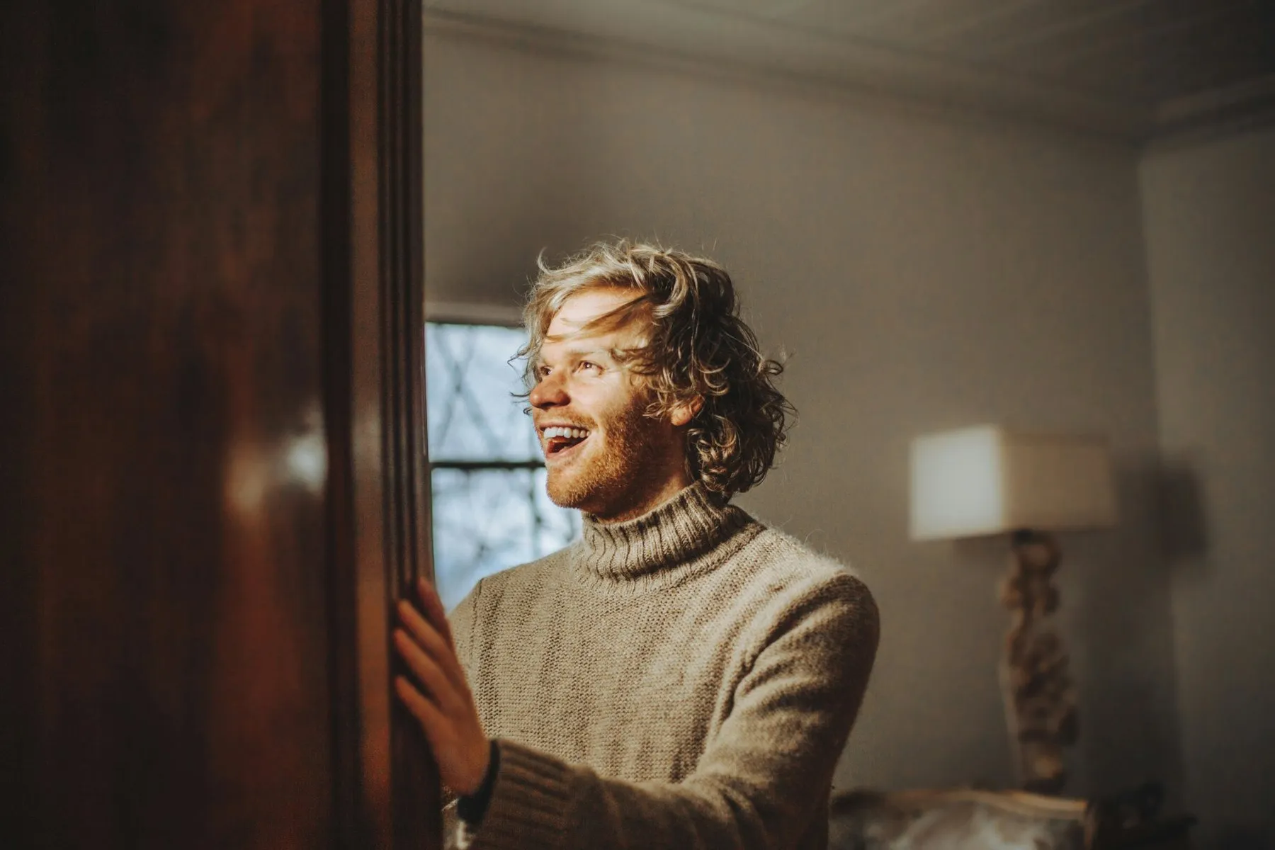 Hombre sonriendo con un jersey de punto grueso de cuello vuelto en tono topo, junto a una puerta de madera