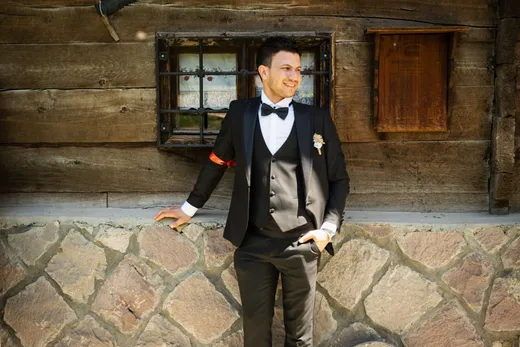 Hombre sonriente con traje negro de tres piezas, camisa blanca, pajarita y flor en la solapa, apoyado en un muro de piedra