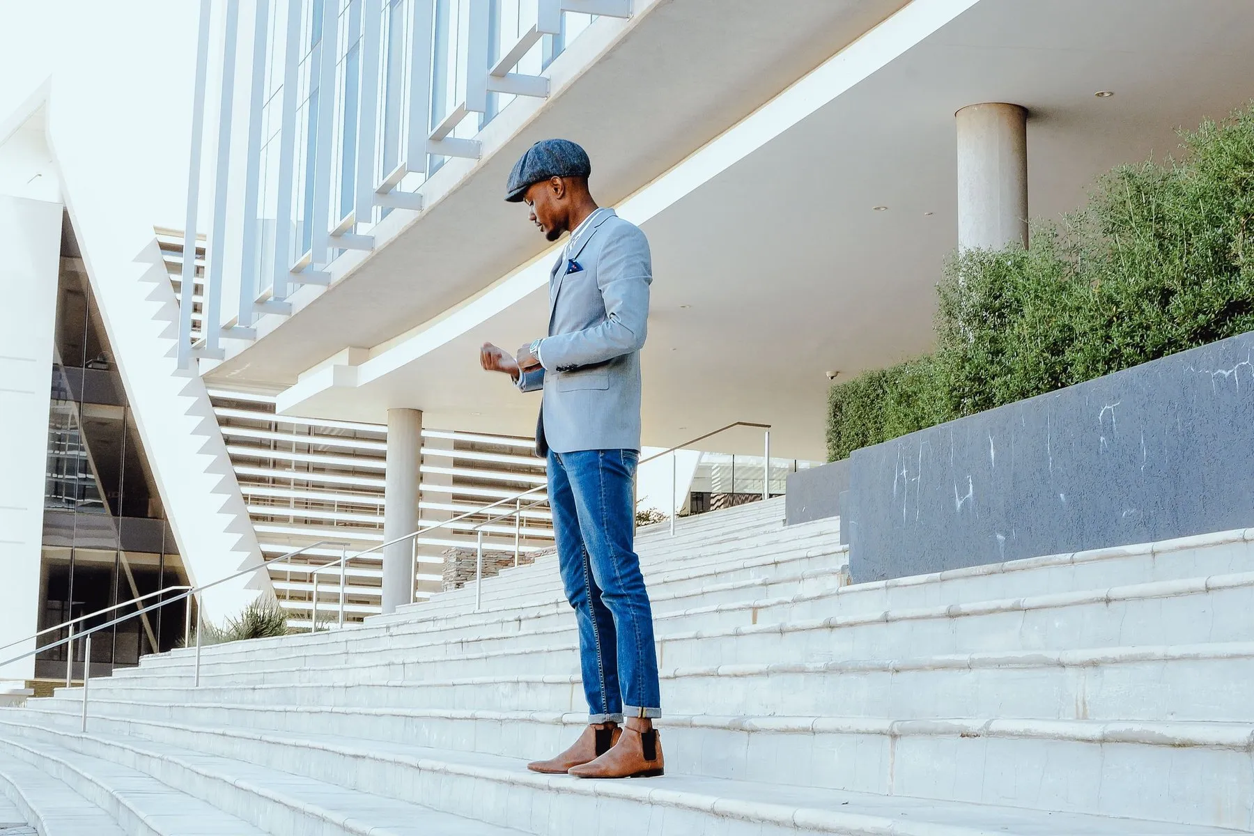 Hombre de perfil en unas escaleras con gorra plana, americana gris azulada, vaqueros azules remangados y botines chelsea marrones